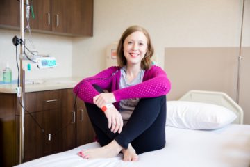 A white woman wearing a bright pink cardigan and black leggings sits with her legs crossed on a hospital bed
