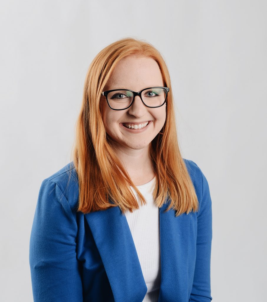 A headshot of Emily Blobaum, who has strawberry blonde hair, blue eyes and glasses. She is wearing a white shirt with a blue blazer.