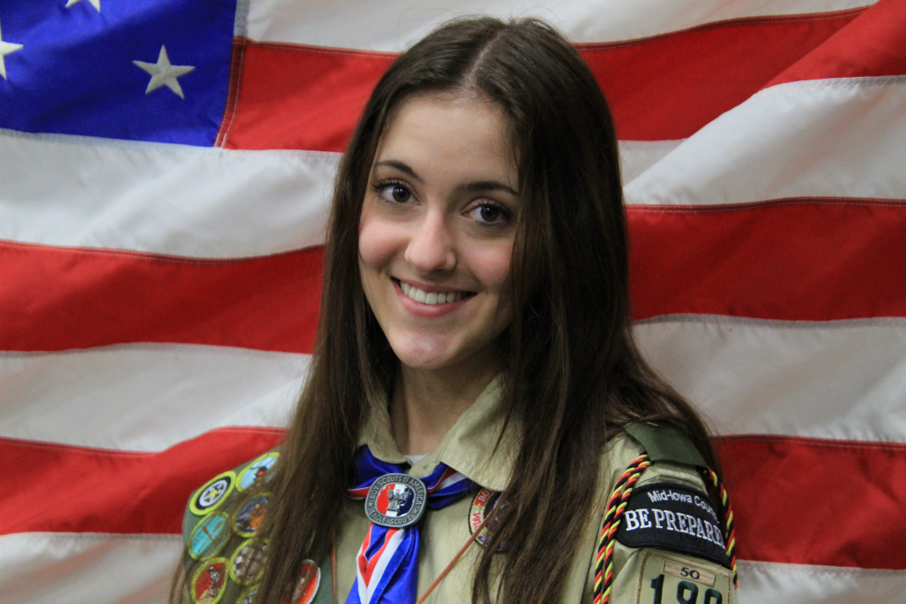 A girl with long brown hair in a khaki Scouts BSA uniform stands in front of an American flag
