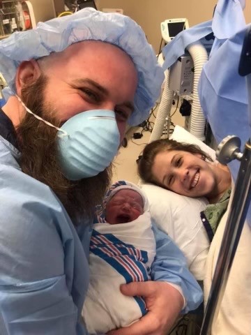 A man with a long brown beard and a blue face mask holds up a newborn baby who is crying. A woman is smiling on a bed in the background