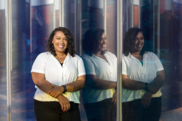A woman with shoulder-length brown curly hair wears a short sleeve white button down shirt. She is posing next to a purple reflective wall, smiling.