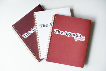 A stack of three planners, one red, one white and one light red. They all say The Agenda Period on them.