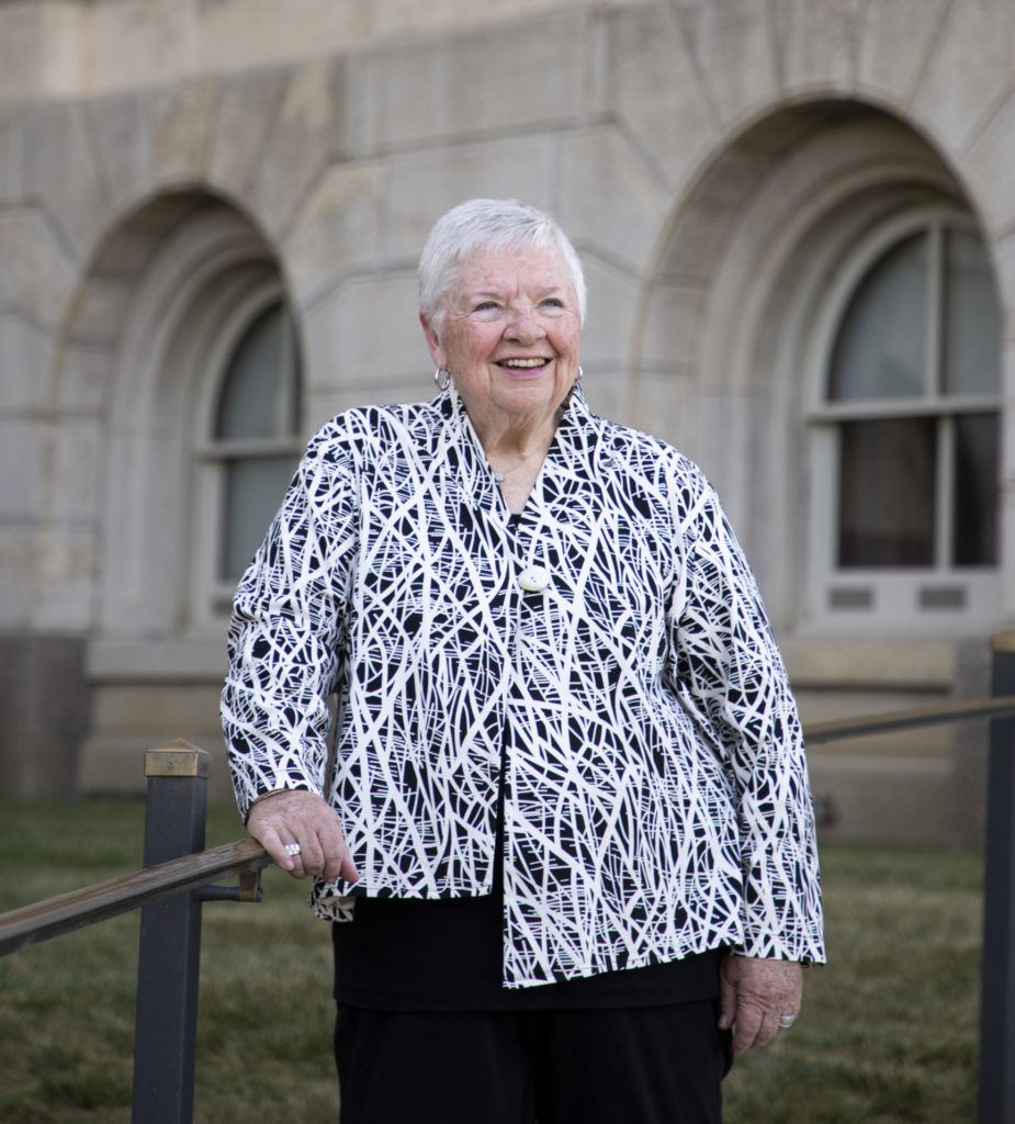 A woman with short white hair wearing a patterned jacket stands in front of the Iowa Capitol building.