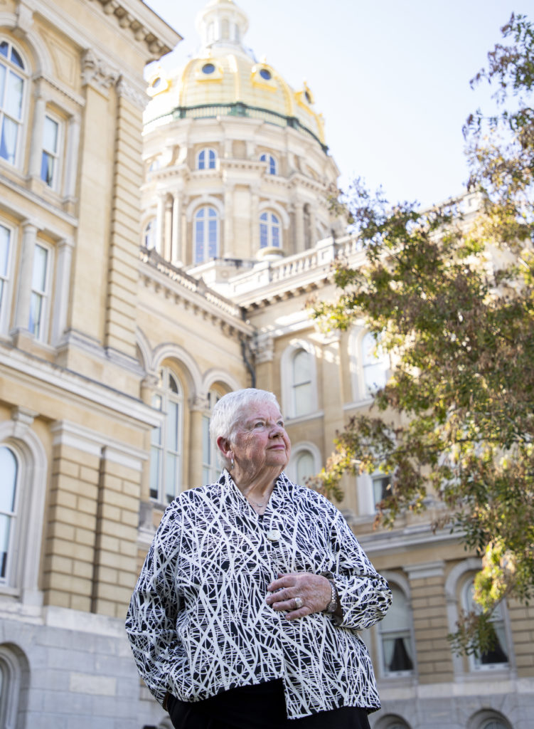 A woman with short white hair wearing a patterned jacket stands in front of the Iowa Capitol building.