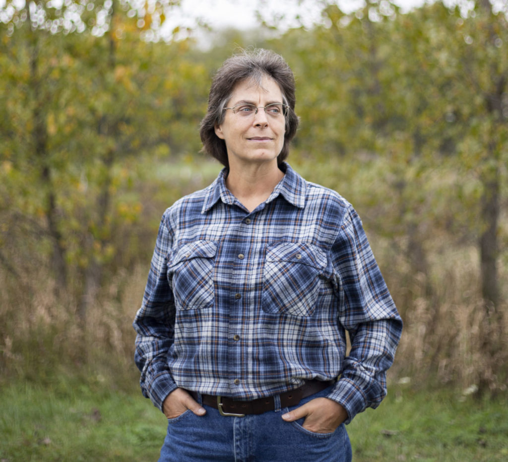 A woman wearing a blue plaid shirt and jeans stands in between two trees and looks away from the camera.