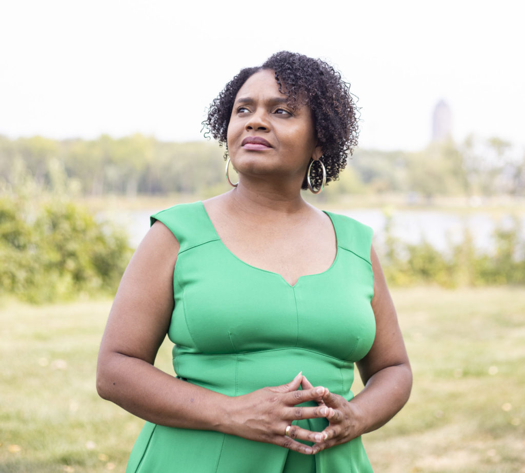 A woman with short curly brown hair wearing a green sleeveless dress looks away from the camera.