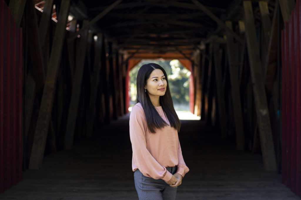 A woman with long dark hair wearing a light pink shirt stands at the entrance of a covered bridge.