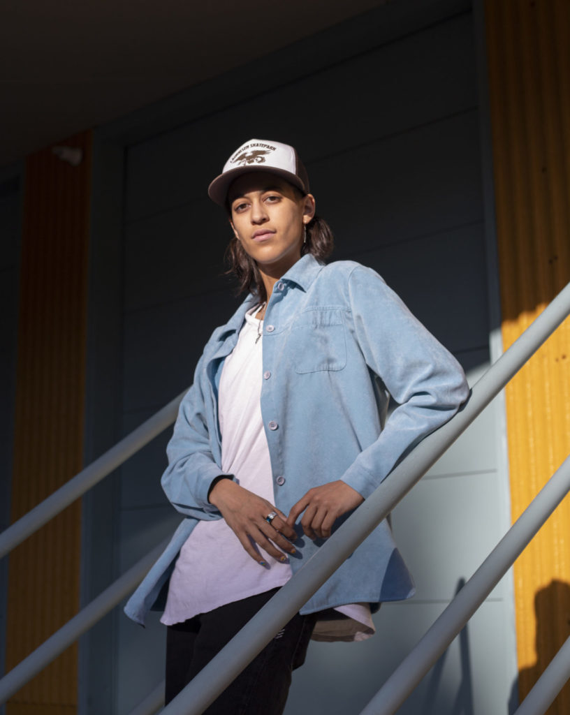 A person wearing a hat and a light blue jacket over a white shirt poses for a photo on a railing.