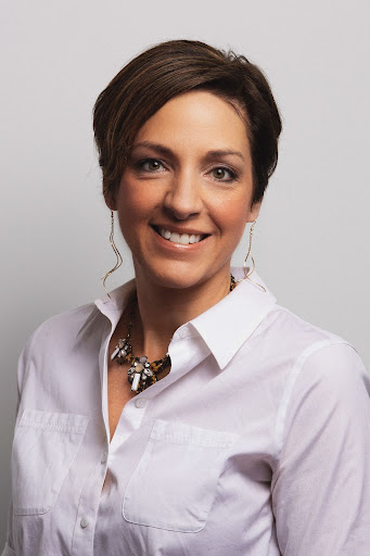 A woman with brown hair wearing a white button-down shirt smiles in front of a white background.