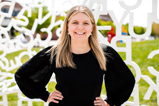 A woman wearing a long sleeve black shirt smiles in front of a sculpture made from white letters.