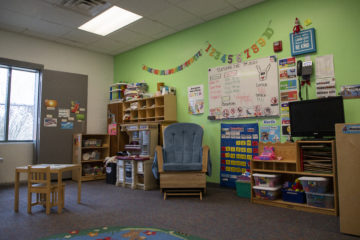 An empty chair sits in the middle of a colorful child care classroom. The walls are green and there are a lot of posters and toys.